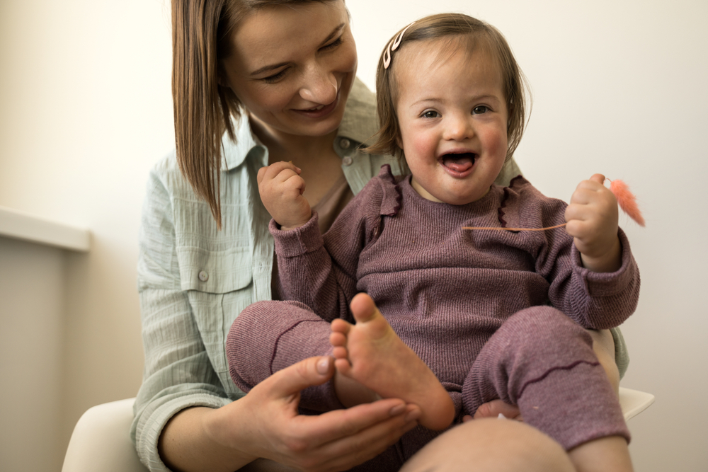 Child with Down's Syndrome aged 18mths smiling and wearing a mauve dungaree outfit whilst having her foot touched by a woman on whose lap the child is sitting. The woman is also smiling