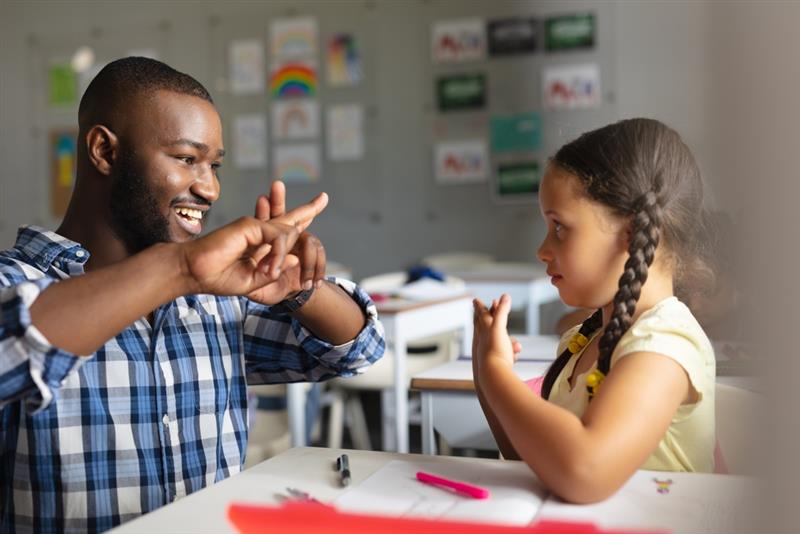 Smiling young male teacher teaching sign language to primary-aged student. 