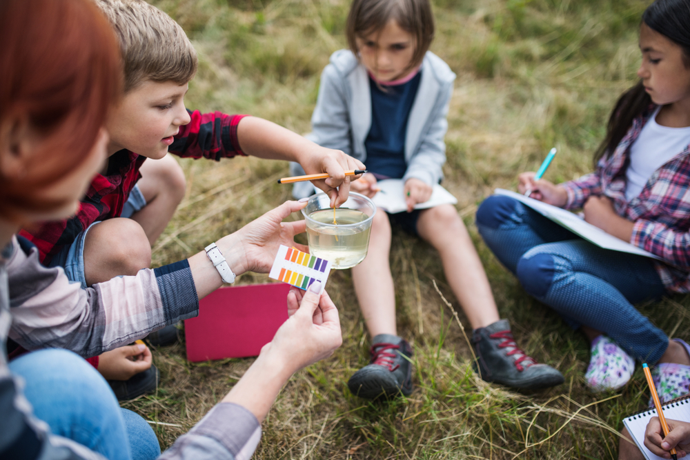 A group of primary-aged students and an adult on a school trip by water with a sample from a river checking pH