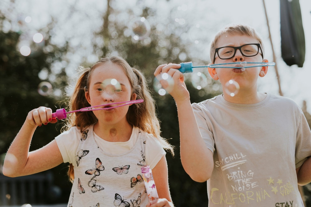 A girl with Down's syndrome and a boy wearing glasses standing side-by-side blowing bubbles 