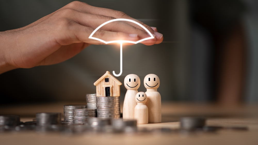 A hand held over piles of coins on a table with wooden figurines and wooden house with a graphic umbrella over the figures