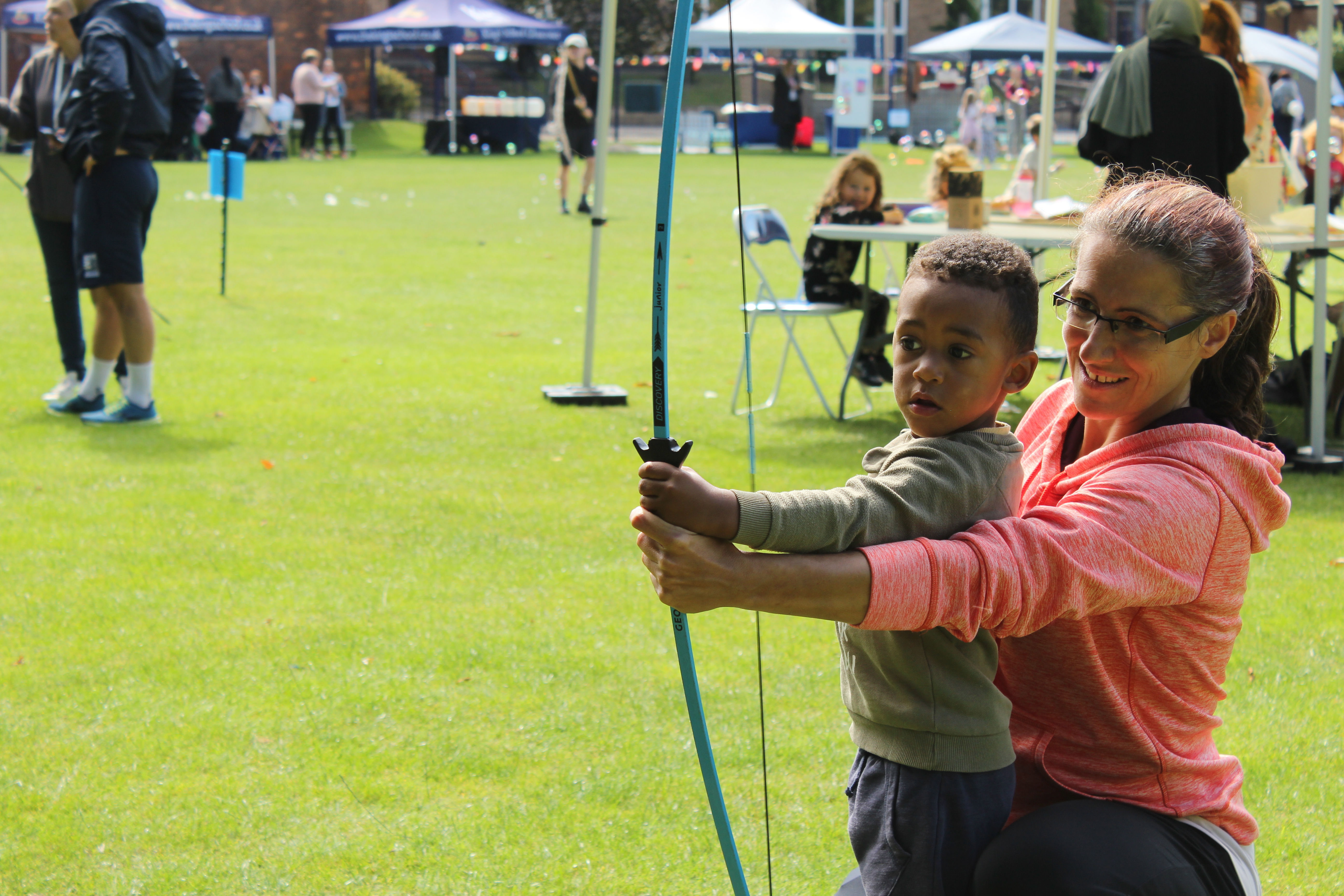 A young boy and a grown up hold a bow and arrow together. They are smiling. This is taking place outdoors at the Gloucestershire 2024 HAF festival at King’s School.