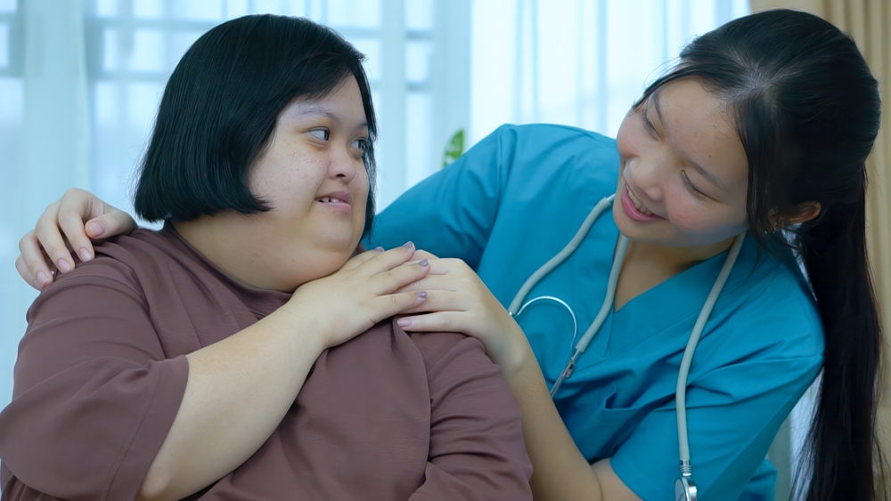Doctor wearing a stethoscope is smiling and supporting patient with Down's Syndrome who is also smiling and they are touching hands