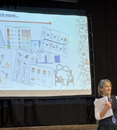 A woman stood in front of a white screen addressing an audience of people sat on chairs.