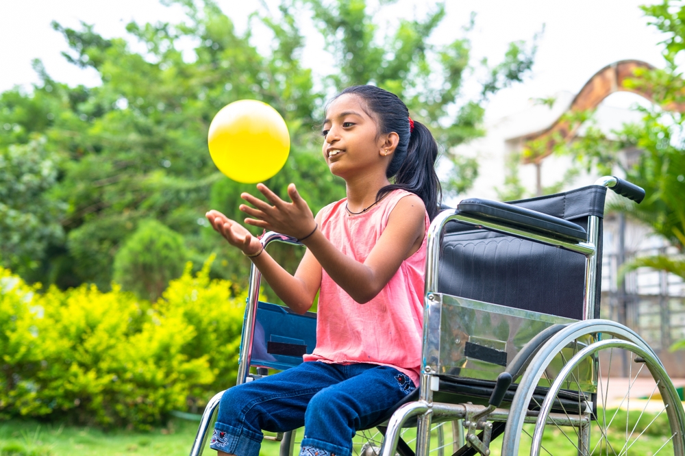 A young girl in a wheelchair smiling and throwing a yellow ball to in the air 