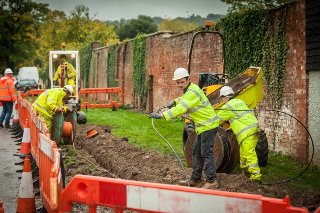 Construction workers in hard hats are laying full fiber cables in a trench separated from the road using orange barriers.