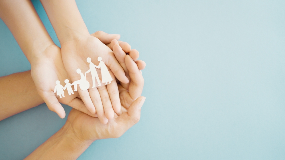 A child's hands in adult hands both holding a paper cut out of an inclusive family