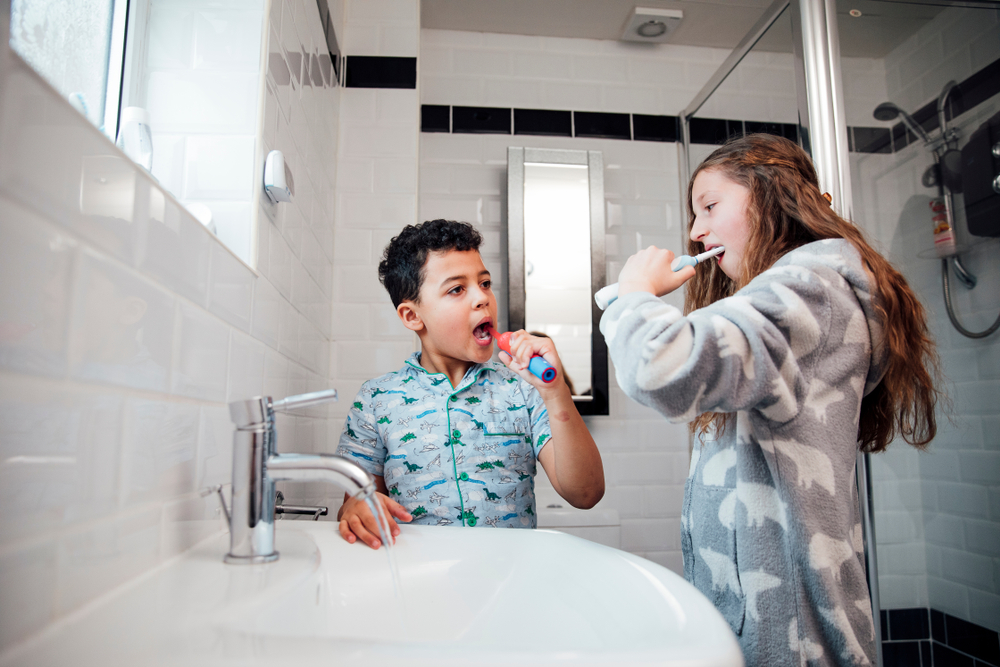 Two children standing in a bathroom using electric toothbrushes