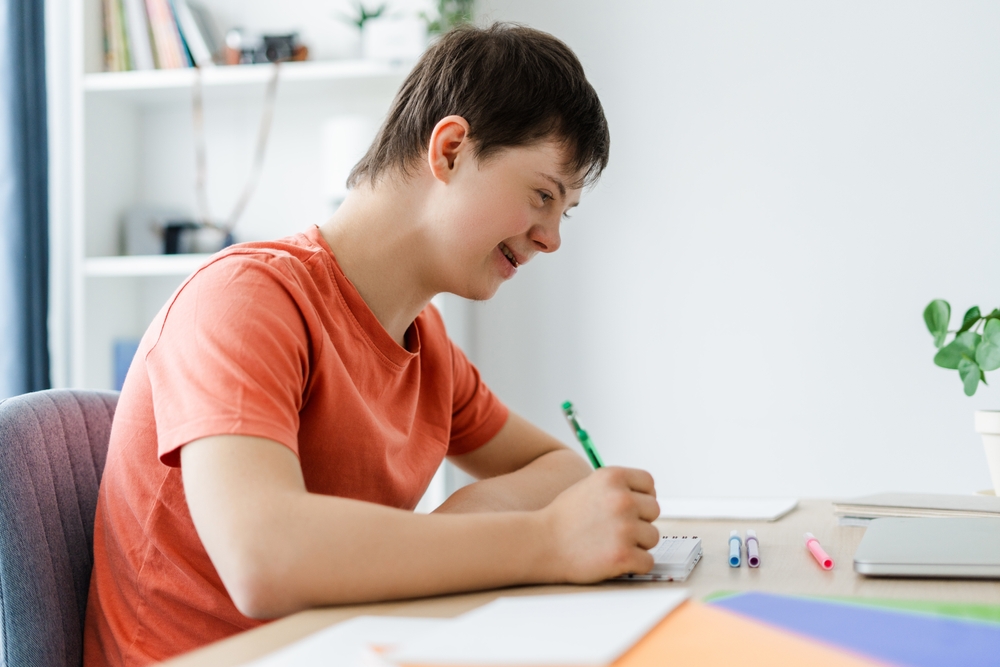  Smiling Young Student With Down Syndrome Is Concentrating Whilst Writing
