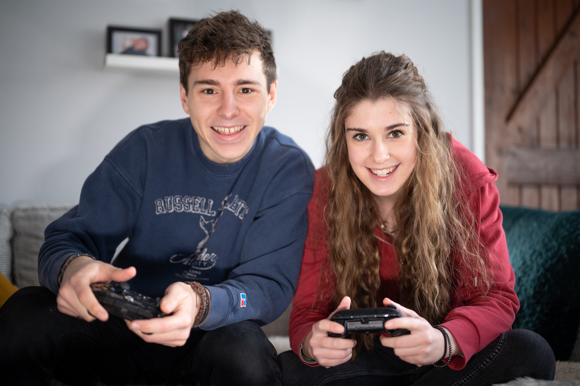 Two people, one wearing a blue sweatshirt and the other a red jacket, sit side by side playing video games with controllers in their hands. Both are smiling and looking towards the camera.