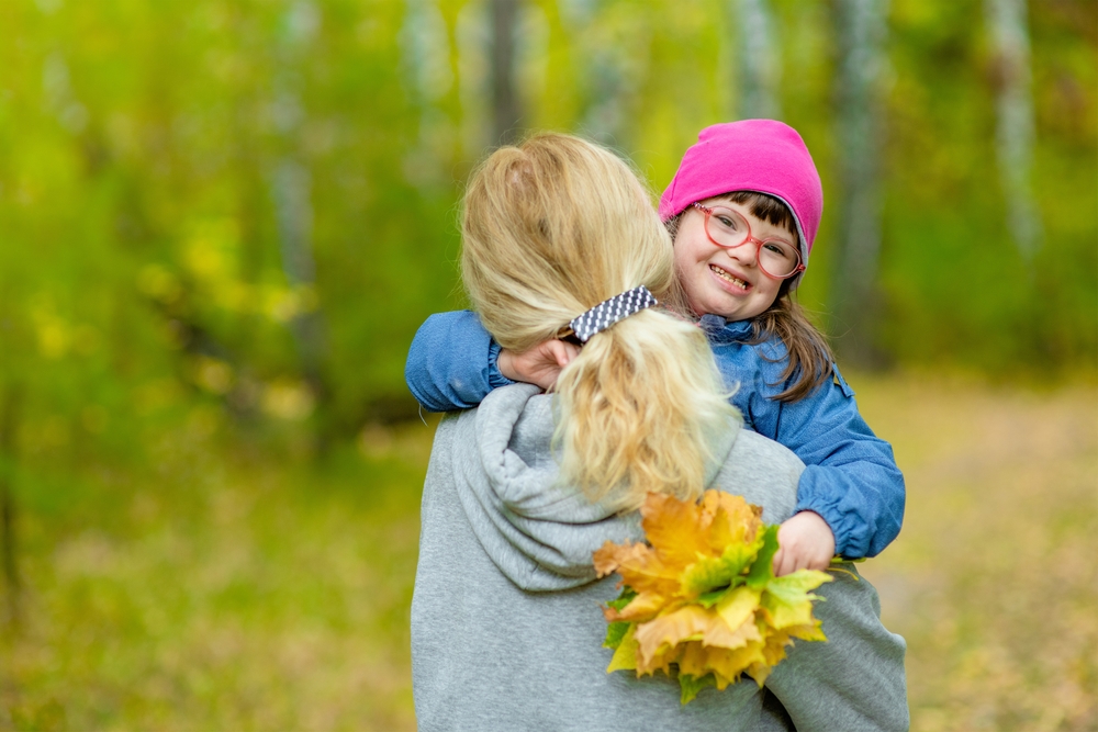 A woman with her back to the camera holding a smiling child whilst walking through a wood in Autumn