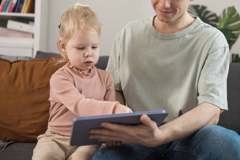 A pre-school child is sitting on a sofa with a man, who is supporting her use of a tablet.