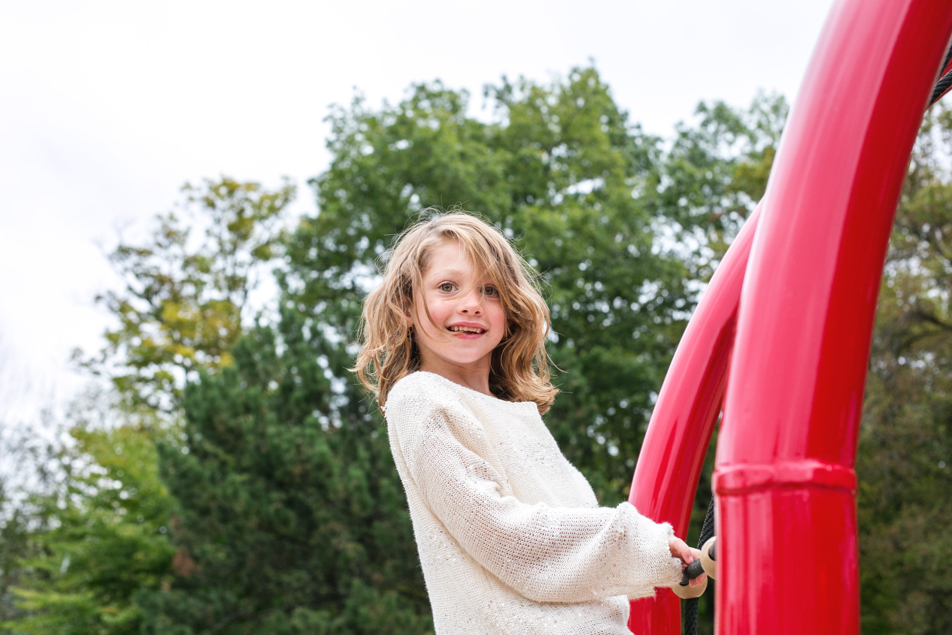 Young girl playing on a red climbing frame, wearing a cream jumper, leaning back and smiling