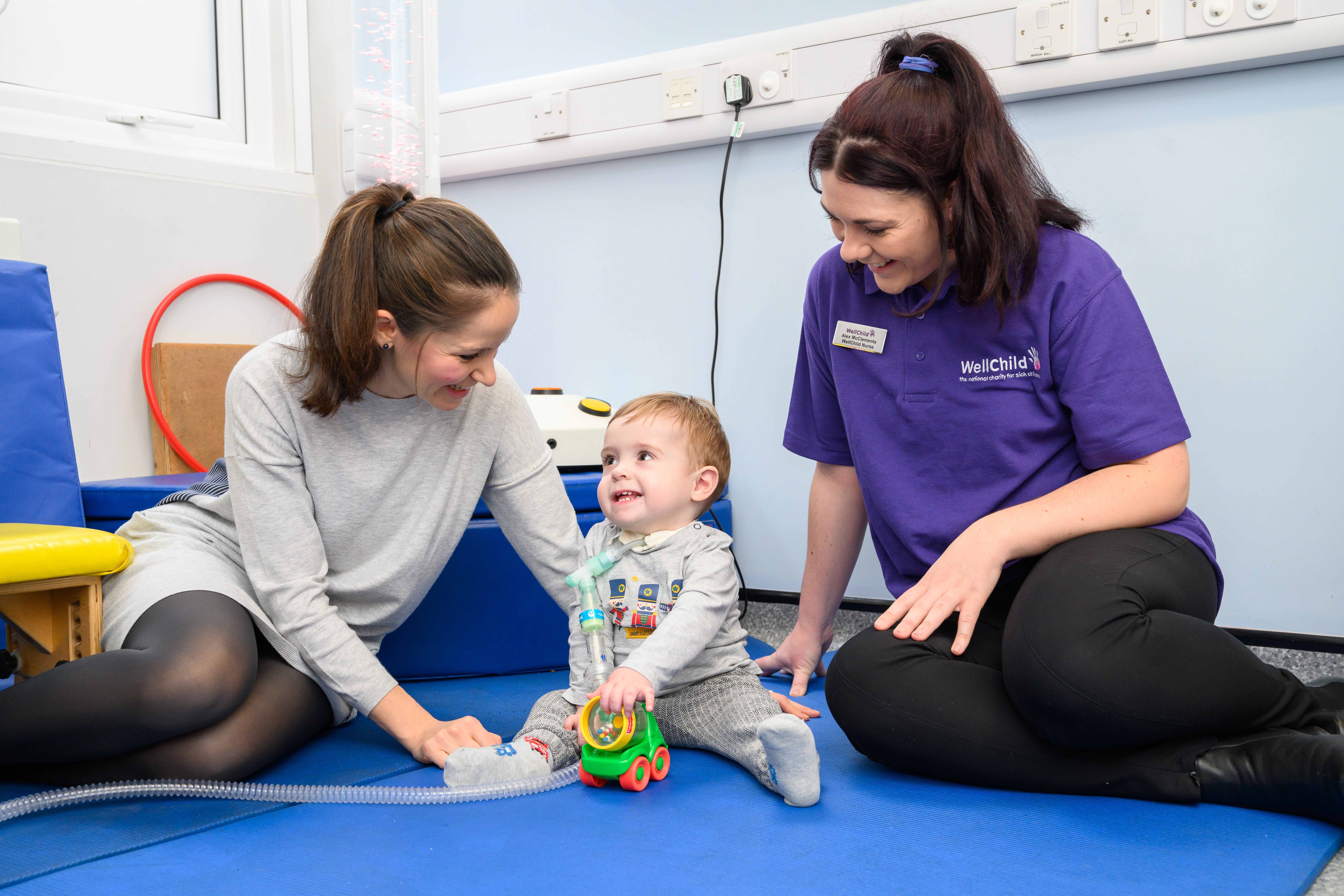 Female WellChild Nurse with long brown hair sits on the floor with a young boy with a tracheostomy and his mum.